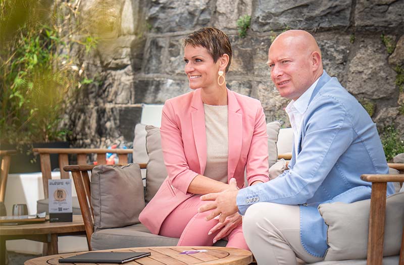 Ein Paar auf einem Sofa sitzend und in ein freundliches Gespräch mit andernen Personen, die nicht sichtbar sind, vertieft